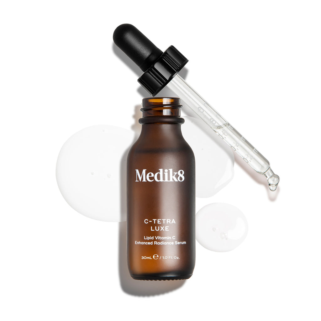 Eine Draufsicht auf eine geöffnete 30-ml-Bernsteinglasflasche des Medik8 C-Tetra Luxe Serums auf weißem Hintergrund. Die Glaspipette liegt neben der Flasche, die von klaren, glänzenden Tropfen des Lipid-Serums umgeben ist.