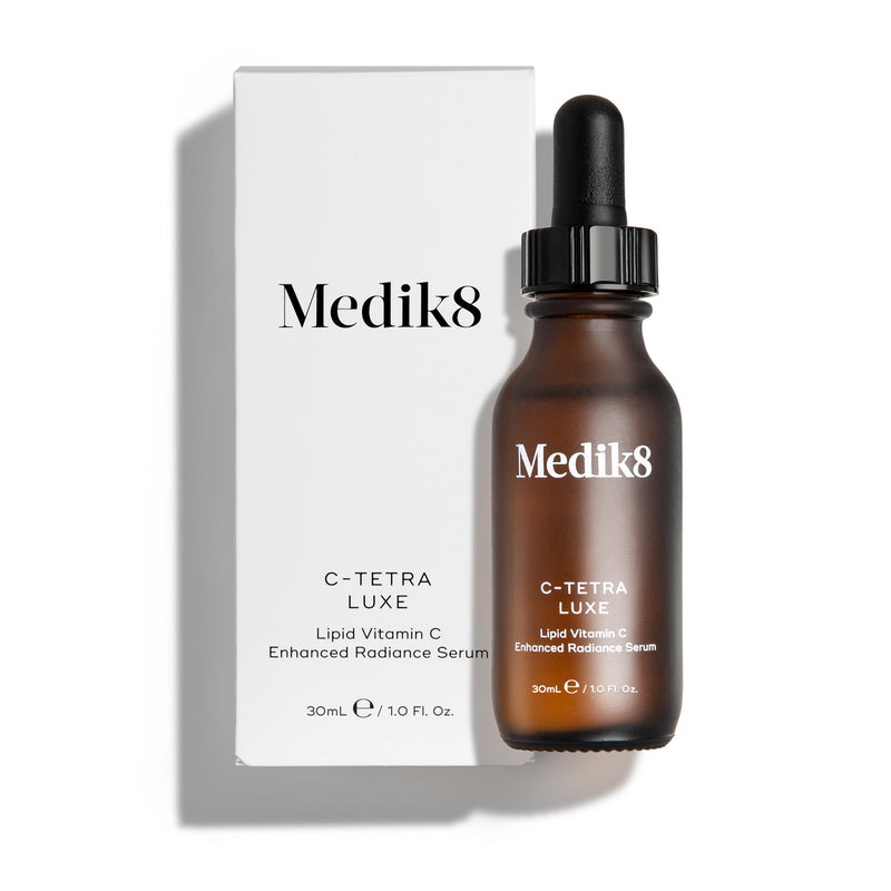 Eine 30-ml-Bernsteinflasche des Medik8 C-Tetra Luxe „Lipid Vitamin C Enhanced Radiance Serum“ neben der weiß-schwarzen Umkarton-Verpackung.