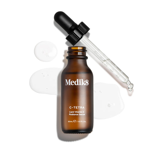 Eine Draufsicht auf eine geöffnete 30-ml-Braunglasflasche des Medik8 C-Tetra Serums. Die Glaspipette liegt daneben, umgeben von klaren, flüssigen Tropfen des Serums auf weißem Hintergrund.