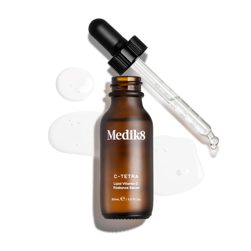 Eine Draufsicht auf eine geöffnete 30-ml-Braunglasflasche des Medik8 C-Tetra Serums. Die Glaspipette liegt daneben, umgeben von klaren, flüssigen Tropfen des Serums auf weißem Hintergrund.