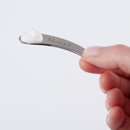 Eine Hand hält einen kleinen, gebogenen silbernen Metallspatel mit dem eingravierten „Medik8“-Logo. Auf der Spitze des Spatels ist ein kleiner Klecks weißer Creme zu sehen.